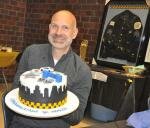 Adam Henkels holds his farewell cake with his flip flop trophy and framed jacket in the background HenkelsWCakeJacket